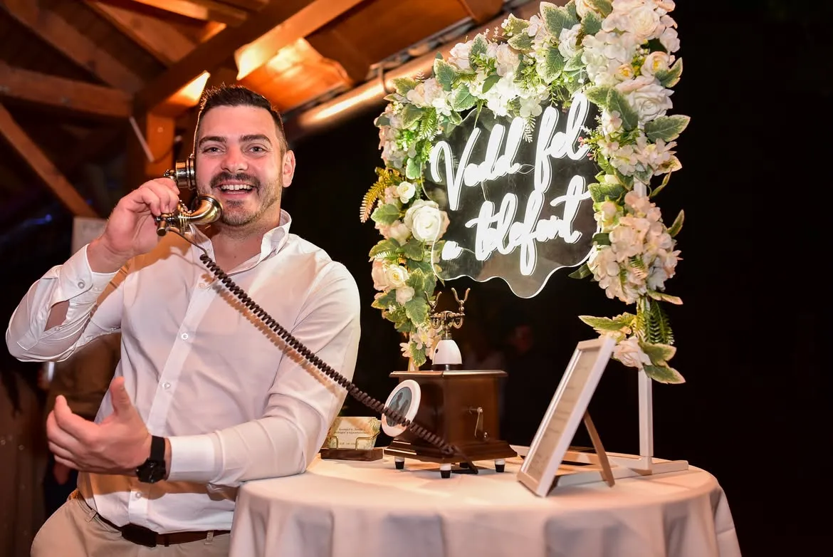 Fehér inges vendég nevetve beszél a ReCall telefonba egy esti rendezvényen, mögötte a fehér rózsákkal keretezett „Vedd fel a telefont” neonfelirat