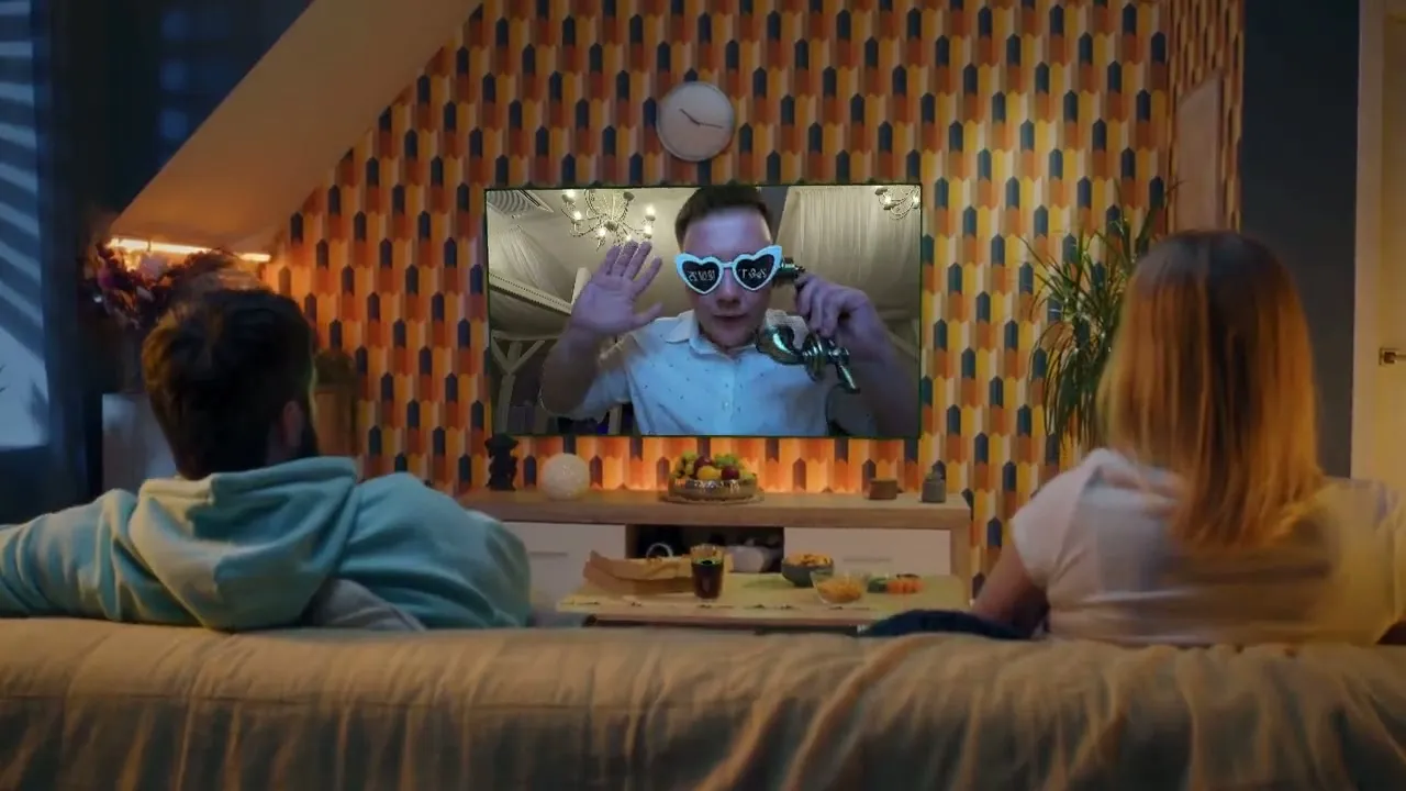 Egy pár a kanapén ülve a tévében nézi vissza a vendégek videóüzeneteit